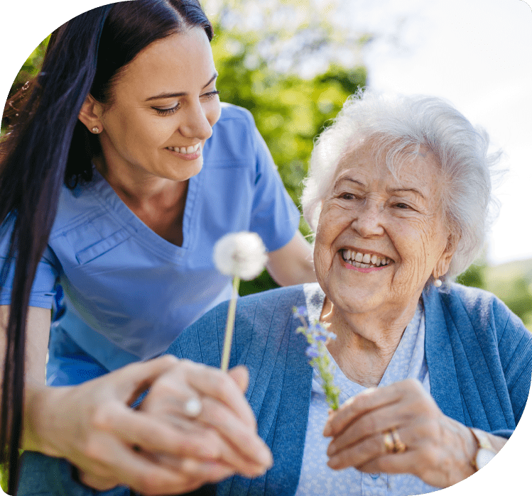Personal Care - Home Care - Specialized Care - Disability Care - Post Hospital Care - Memory care - Dementia care - Companion  Care - 24/7 care - A Rosebuds' caregiver helping an elderly woman with flowers outdoors, both smiling.