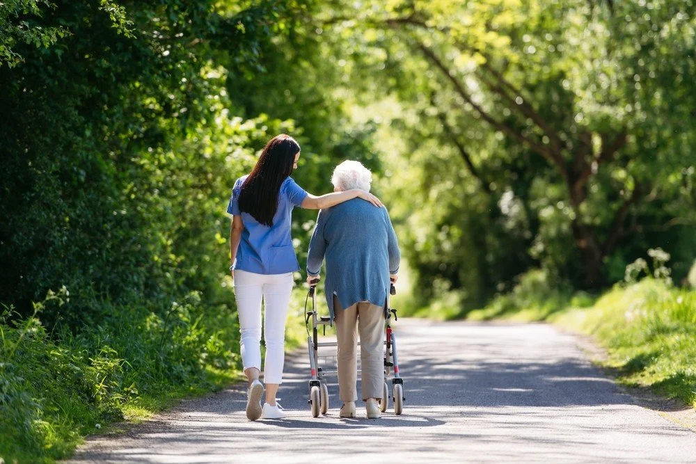 Personal Care - Home Care - Specialized Care - Disability Care - Post Hospital Care - Memory care - Dementia care - Companion  Care - 24/7 care - A Rosebuds' caregiver helping an elderly woman walk with a walker in a lush green park.