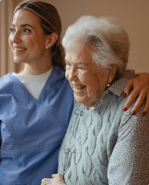 Personal Care - Home Care - Specialized Care - Disability Care - Post Hospital Care - Memory care - Companion Care - A young female Rosebuds' caregiver embraces an elderly woman with white hair, who is smiling and looking off to the side.