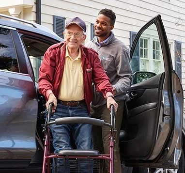 Personal Care - Home Care - Specialized Care - Disability Care - Post Hospital Care - Memory care - Dementia care - Companion Care - 24/7 care - An elderly man using a walker being assisted into a car by a Rosebuds' caregiver outside a house.