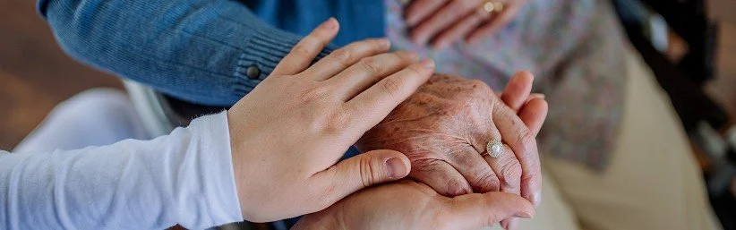 Personal Care - Home Care - Specialized Care - Disability Care - Post Hospital Care - Memory care - Companion care - A Rosebuds' caregiver's hand resting on an elderly person's bent knee, with another hand gently holding the elderly person's hand.