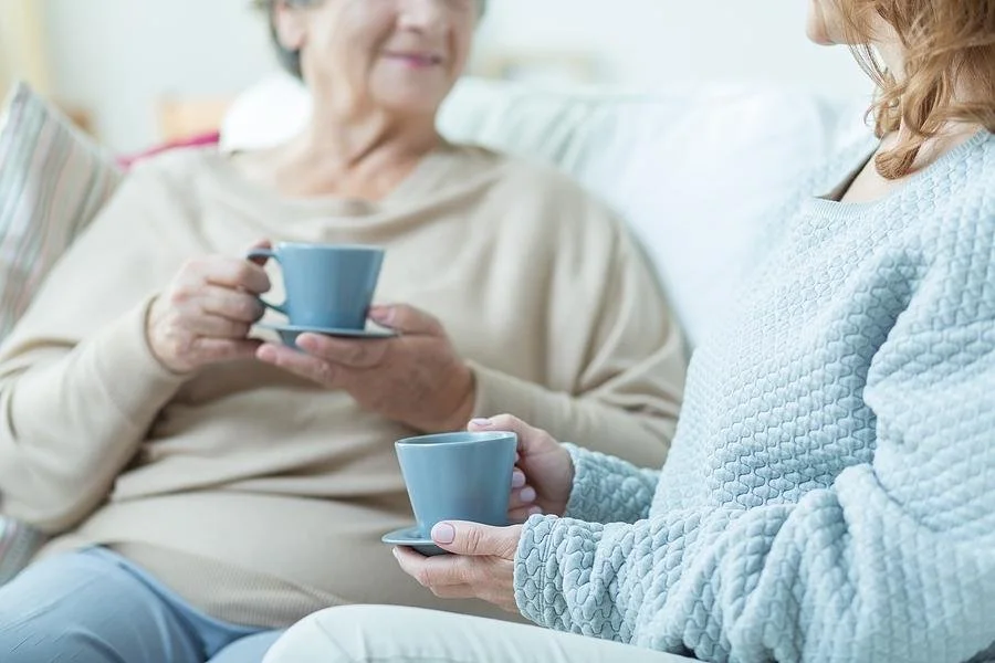 Personal Care - Home Care - Specialized Care - Disability Care - Post Hospital Care - Memory care - Dementia care - Companion  Care - A Rosebuds' caregiver and a senior lady sitting on a sofa, sharing a conversation while holding blue coffee cups.