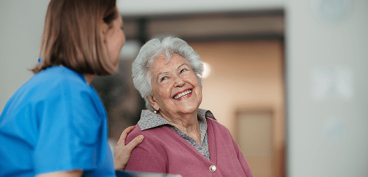 Personal Care - Home Care - Specialized Care - Disability Care - Post Hospital Care - Memory care - Dementia care - Companion  Care - An elderly woman smiling and looking at a caregiver who is gently touching her shoulder in a home.