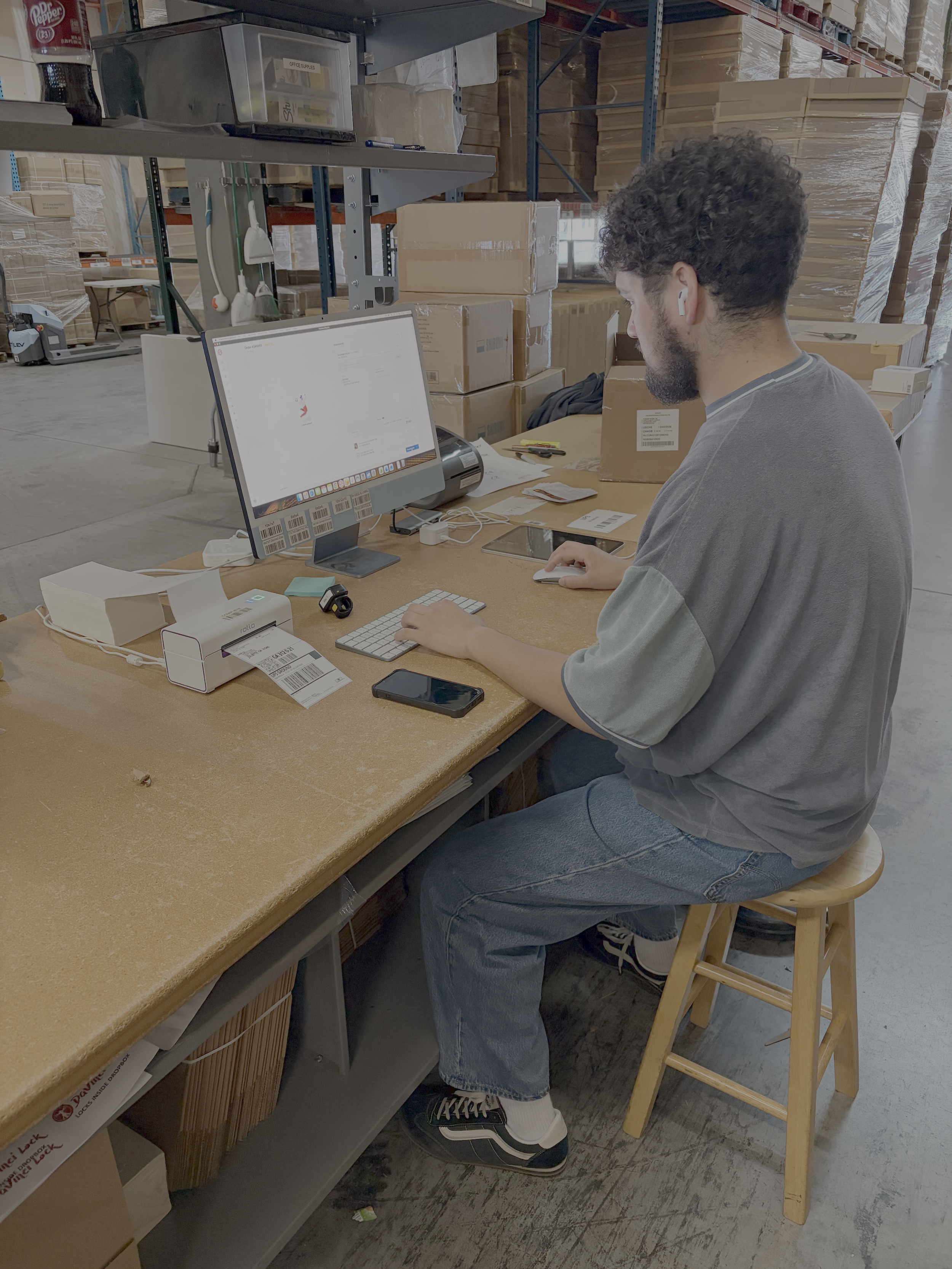 A man working at a desk in a warehouse, using a computer with a barcode scanner nearby, surrounded by cardboard boxes and warehouse shelves.