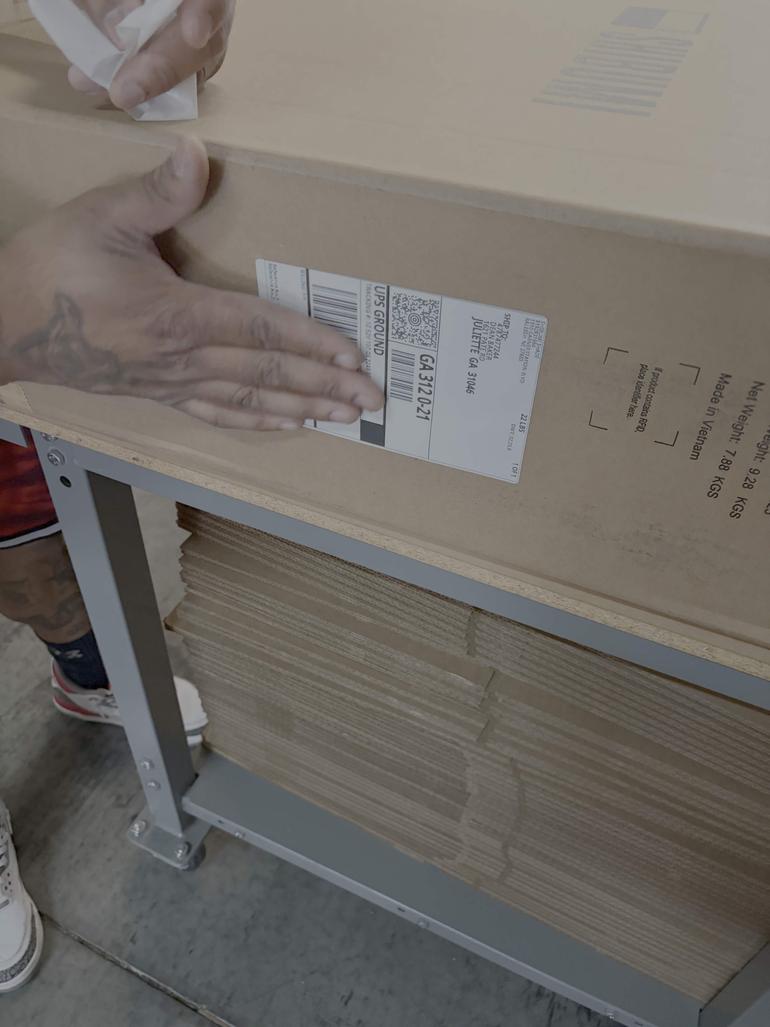 Person placing a shipping label on a cardboard box in a warehouse or store, with additional stacked boxes on a metal shelf below.