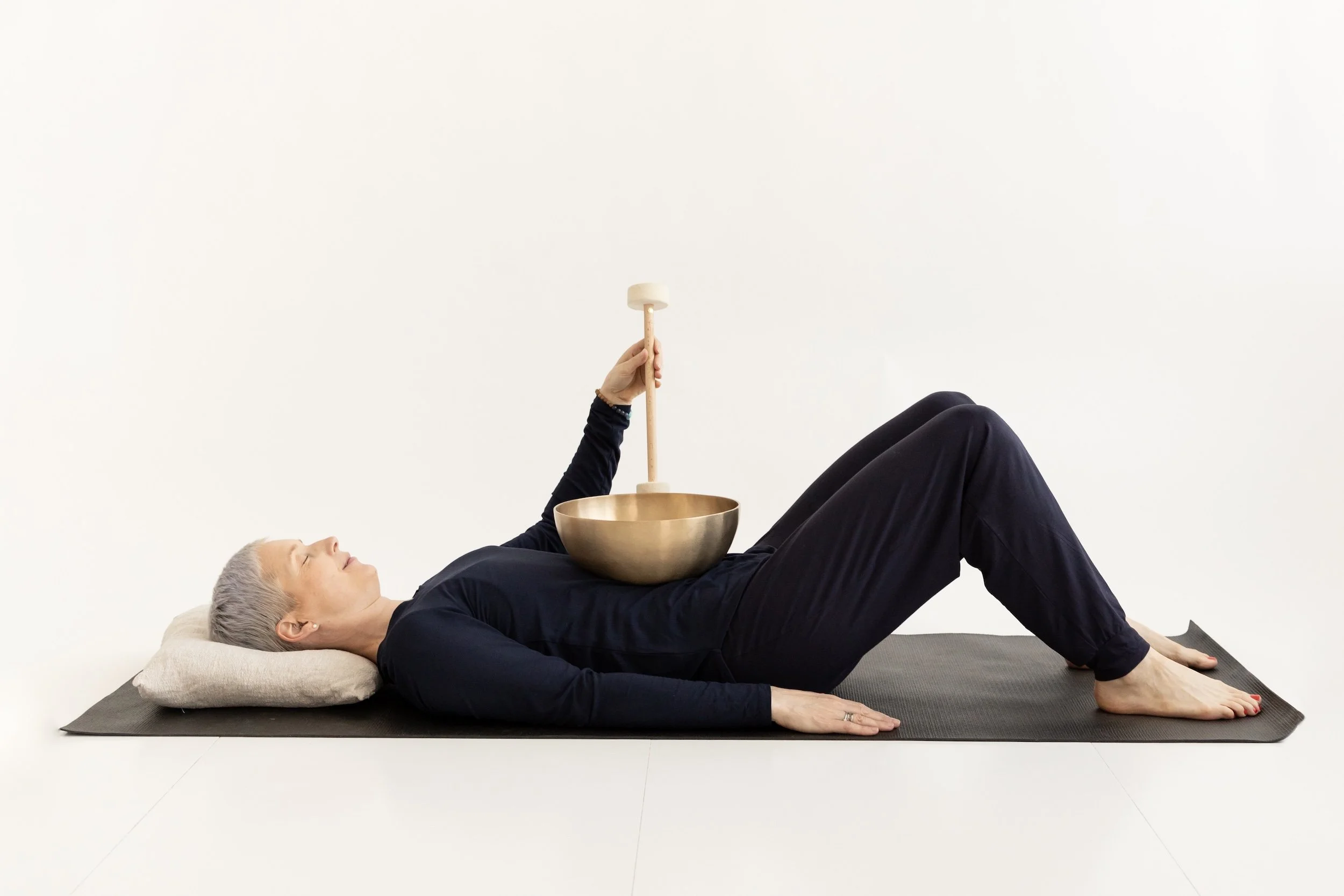 Ihminen makaa selällään matolla äänimalja vatsan päällä, pitäen käsivartensa auki ja silmänsä kiinni. Hän käyttää tummansinistä pitkähihaista yläosaa ja mustia housuja. Hänen päänsä lepää tyynyllä, ja hän rentoutuu äänimaljarentoutuksessa.