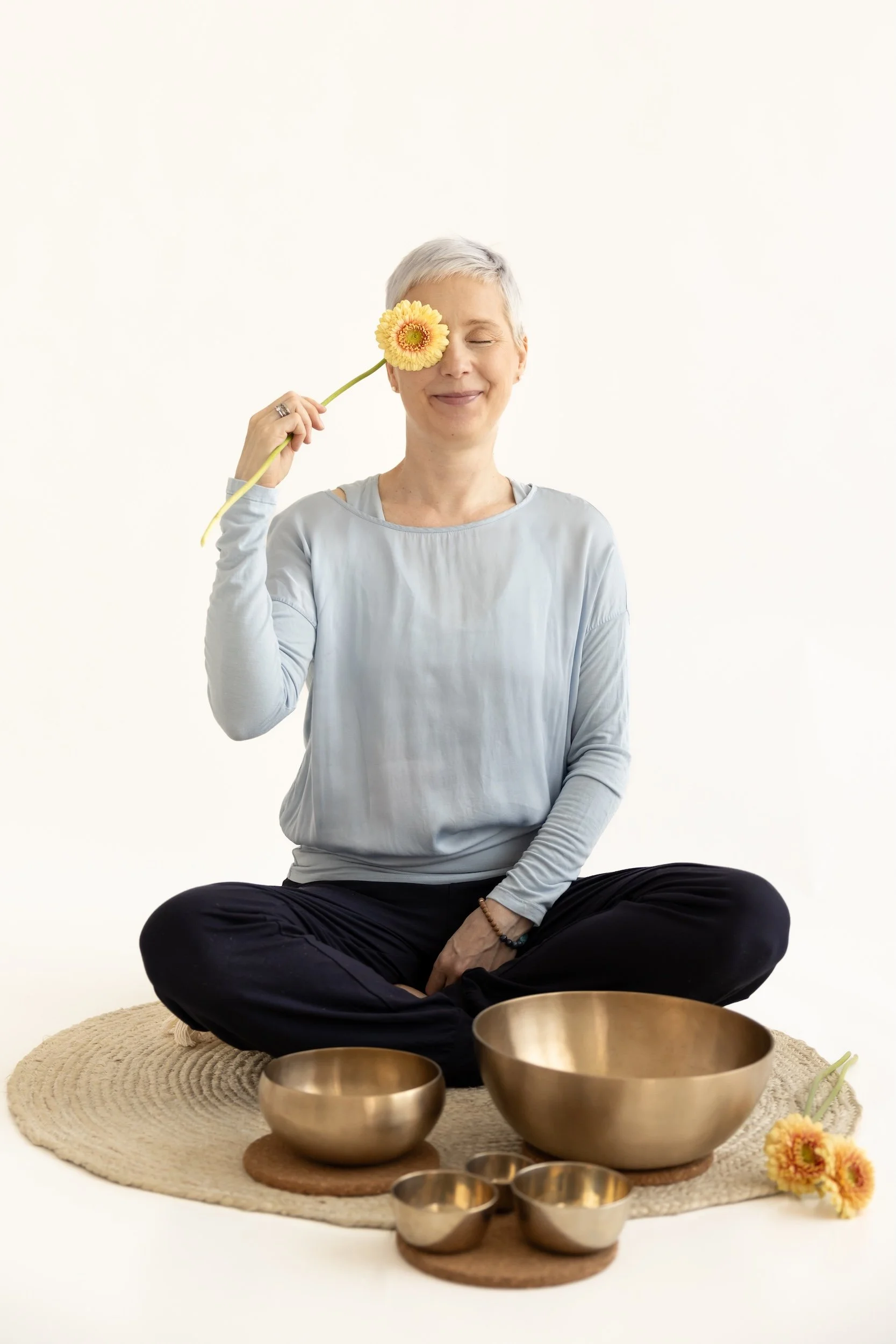 Nainen istuu äänimaljojen takana matolla, ympärillä metallisia maljoja ja kukkia, yksi kukka on silmien edessä.