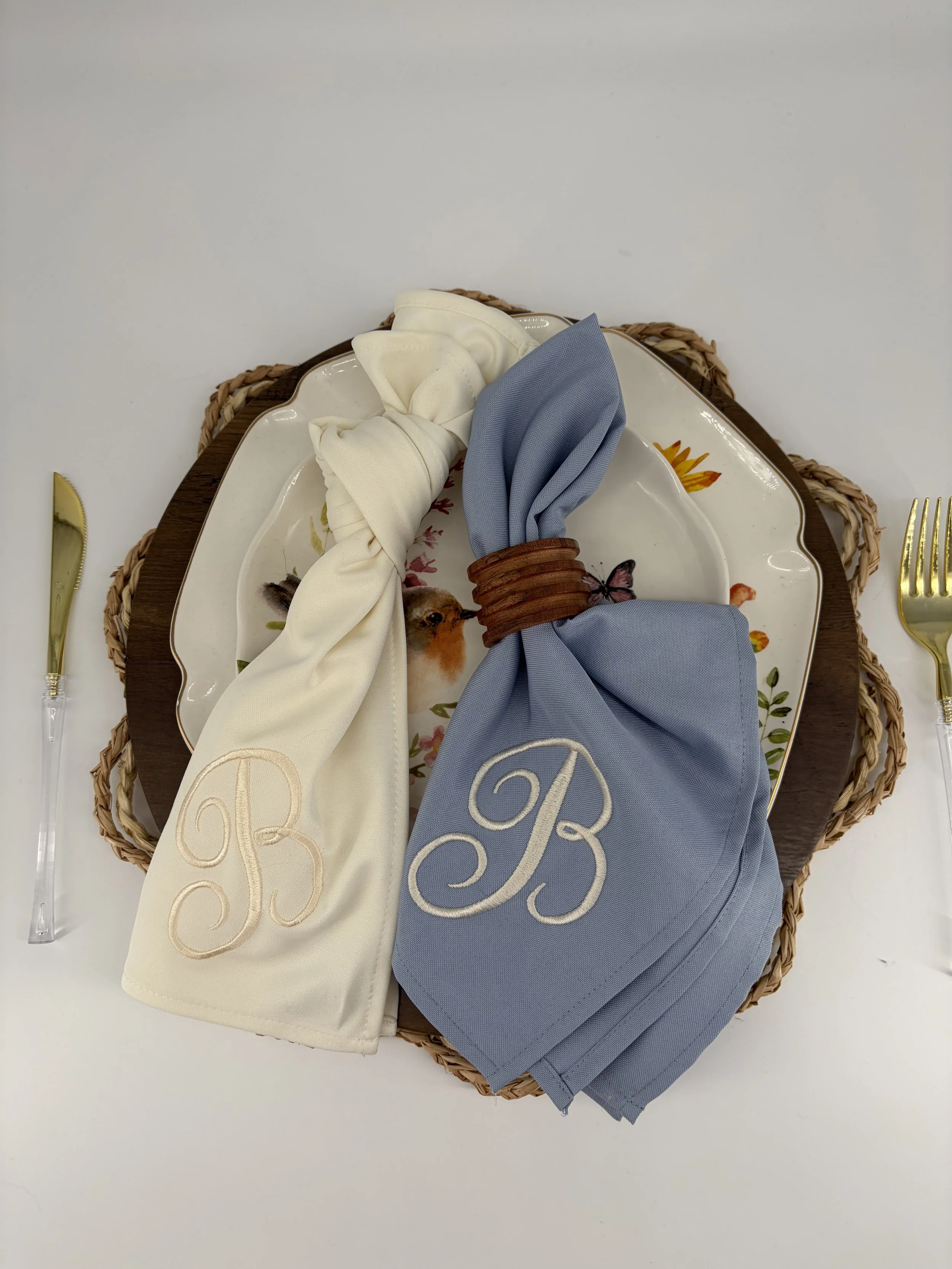 A table setting with a straw placemat, a floral-patterned plate, and two folded cloth napkins in cream and blue, with monogrammed initials, placed on the plate.
