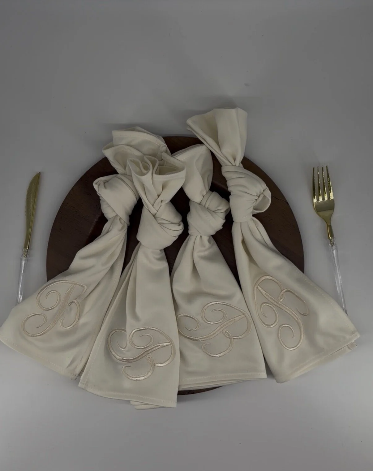 Elegant table setting with four knotted cream-colored napkins with embroidered initials, gold-handled knife and fork, and a wooden round platter on a white tablecloth.