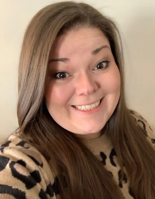 A woman with long light brown hair smiling at the camera, wearing a beige top with a black leopard print pattern.