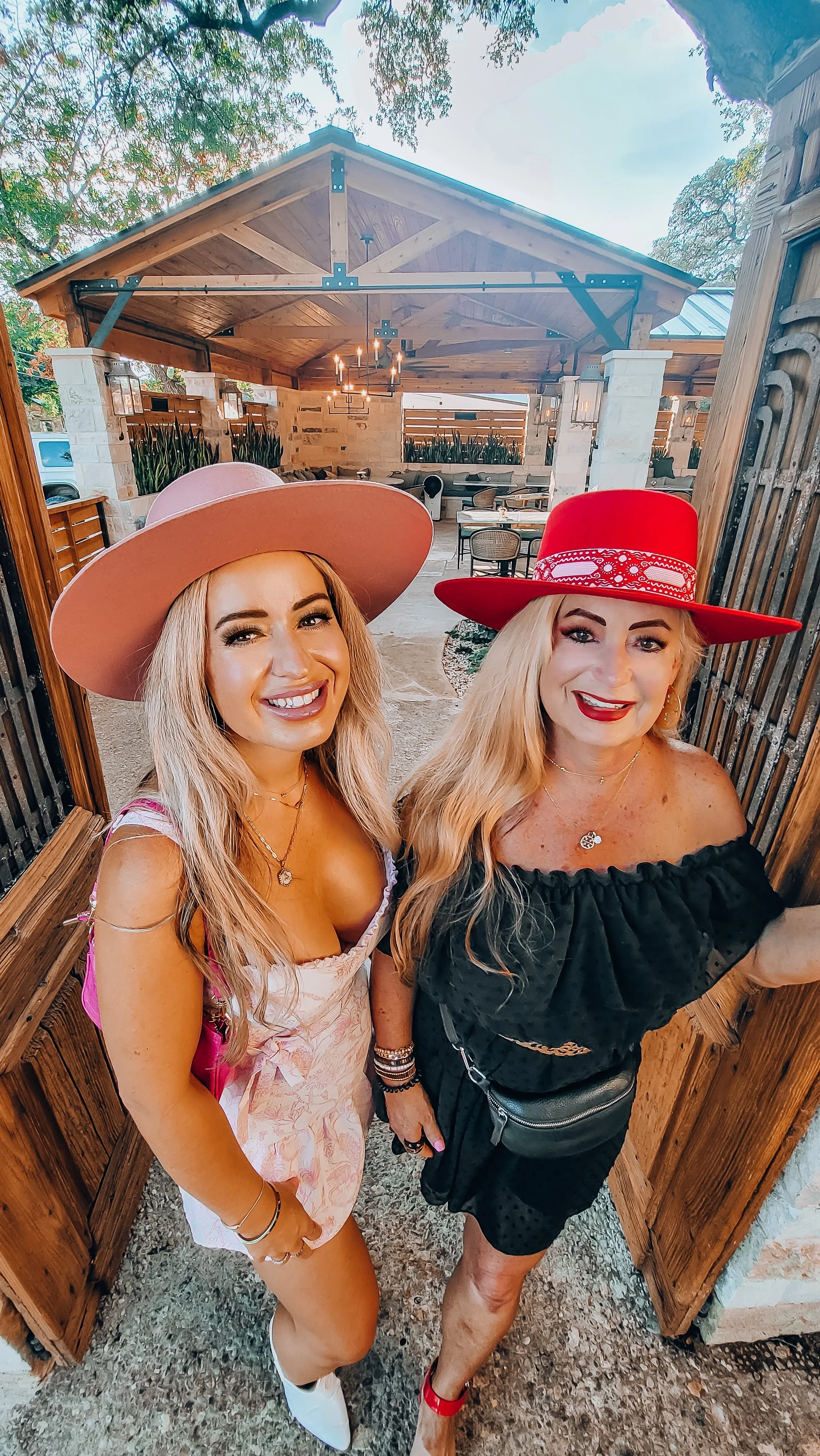 Two women wearing large hats and smiling pose for a photo in an outdoor patio area with a wooden gazebo and decorated with plants and hanging lights.