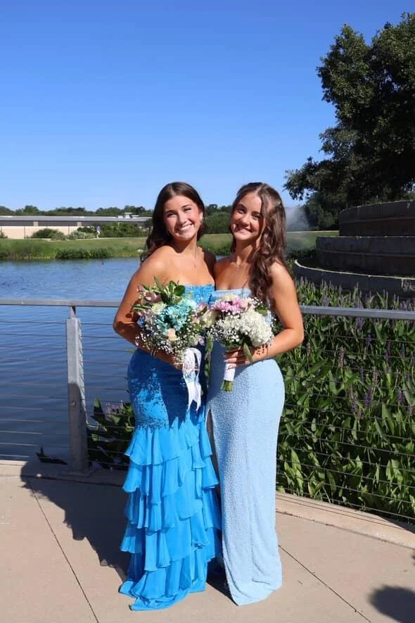 Two young women in blue dresses holding bouquets of flowers, standing outdoors by a body of water with a fountain and greenery in the background.