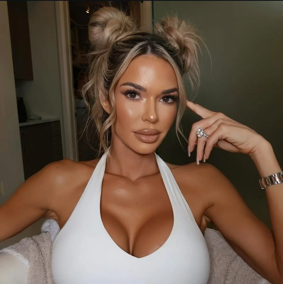 A woman with styled blonde hair in two buns, wearing a white top, posing with her left hand near her face, displaying a ring and a watch, looking into the camera.