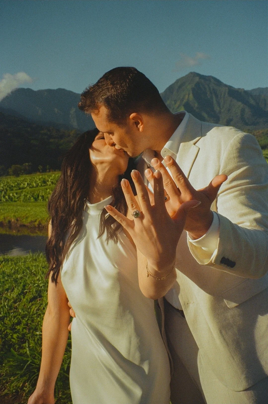 A couple sharing a kiss outdoors with mountains and greenery in the background.