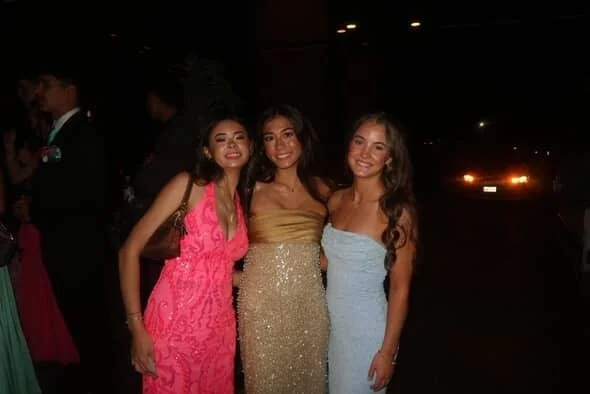 Three young women in colorful dresses posing together at night in an outdoor setting, smiling.