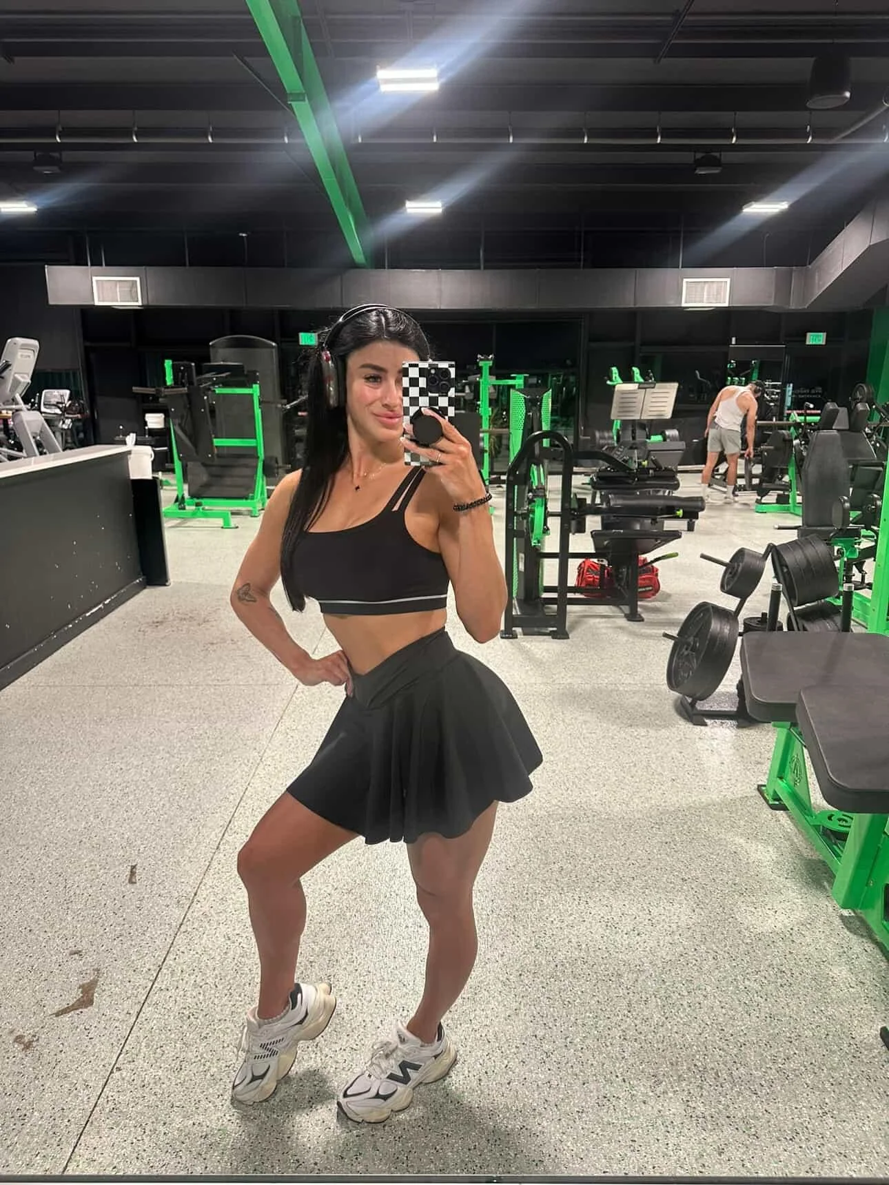 A woman in workout clothes taking a selfie in a gym, wearing a black sports bra, black skirt, white sneakers, and large headphones.