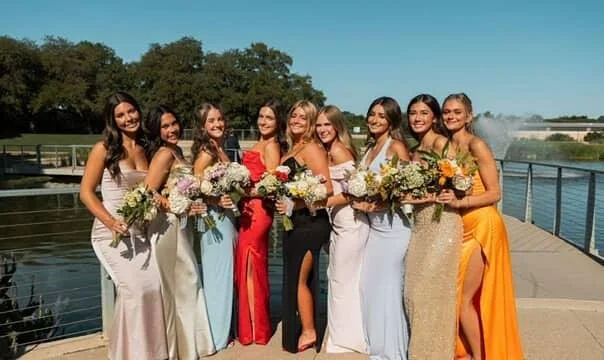 Nine women in formal dresses standing together on a bridge by a river, holding bouquets of flowers, smiling for a group photo.