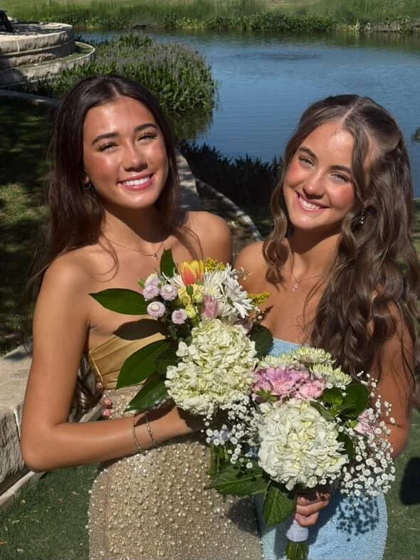 Two women smiling and holding bouquets of flowers near a river, with greenery and a stone structure in the background.