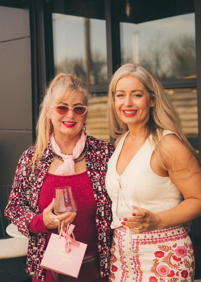 Two women smiling and holding glasses, standing outside in front of a building.