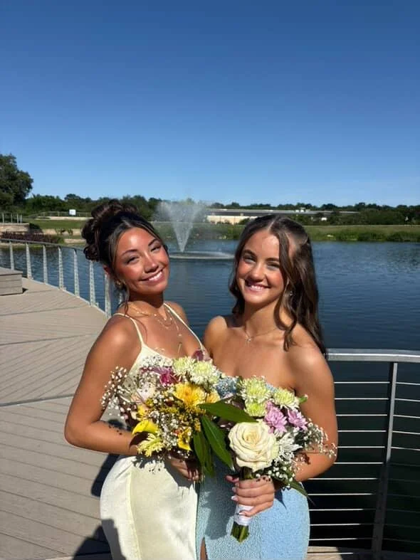 Two young women smiling and holding bouquets of flowers outdoors next to a river with a fountain, under a clear blue sky.