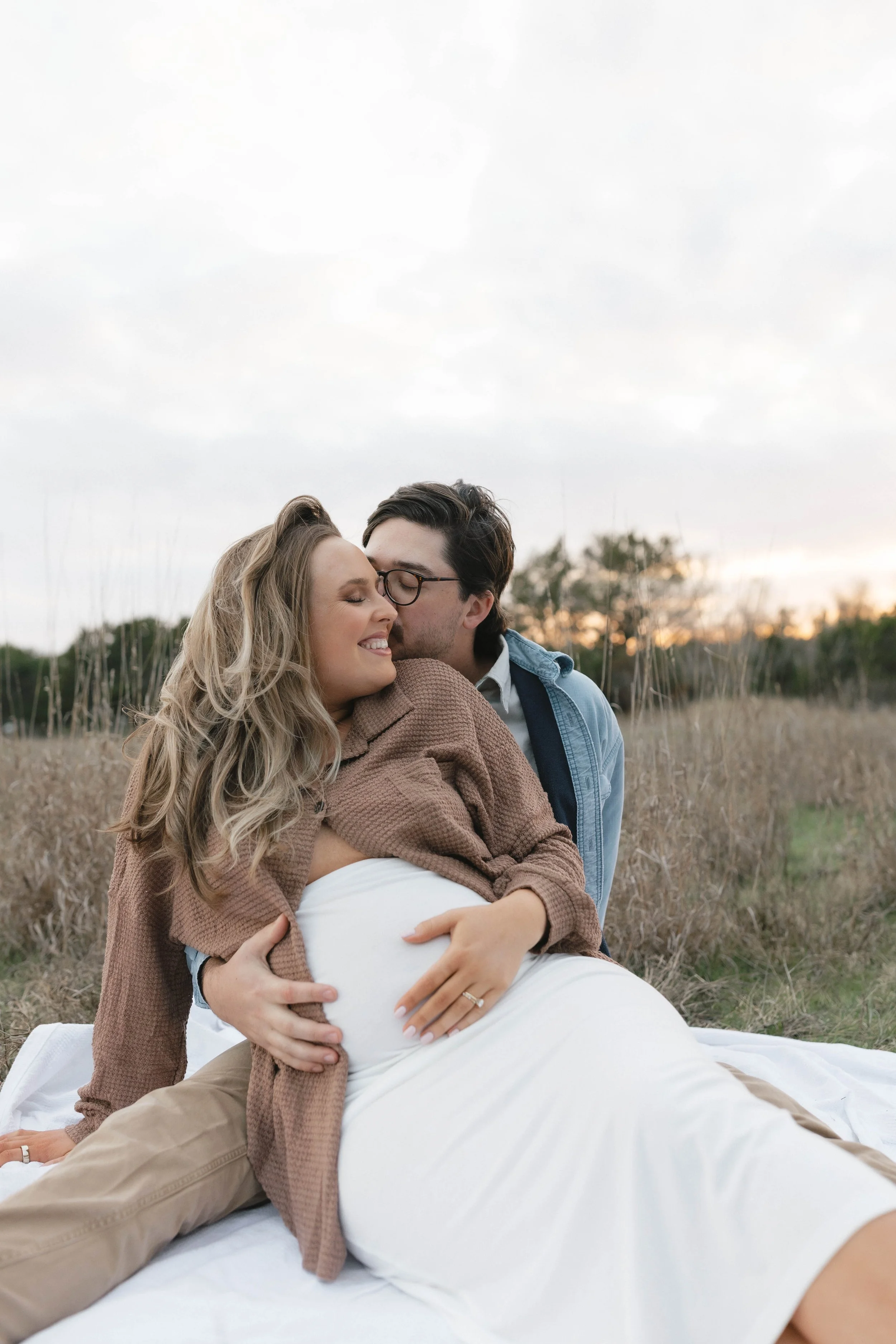 A couple celebrating pregnancy outdoors at sunset, with the man kissing the woman's temple as she smiles, sitting on a blanket in a field.