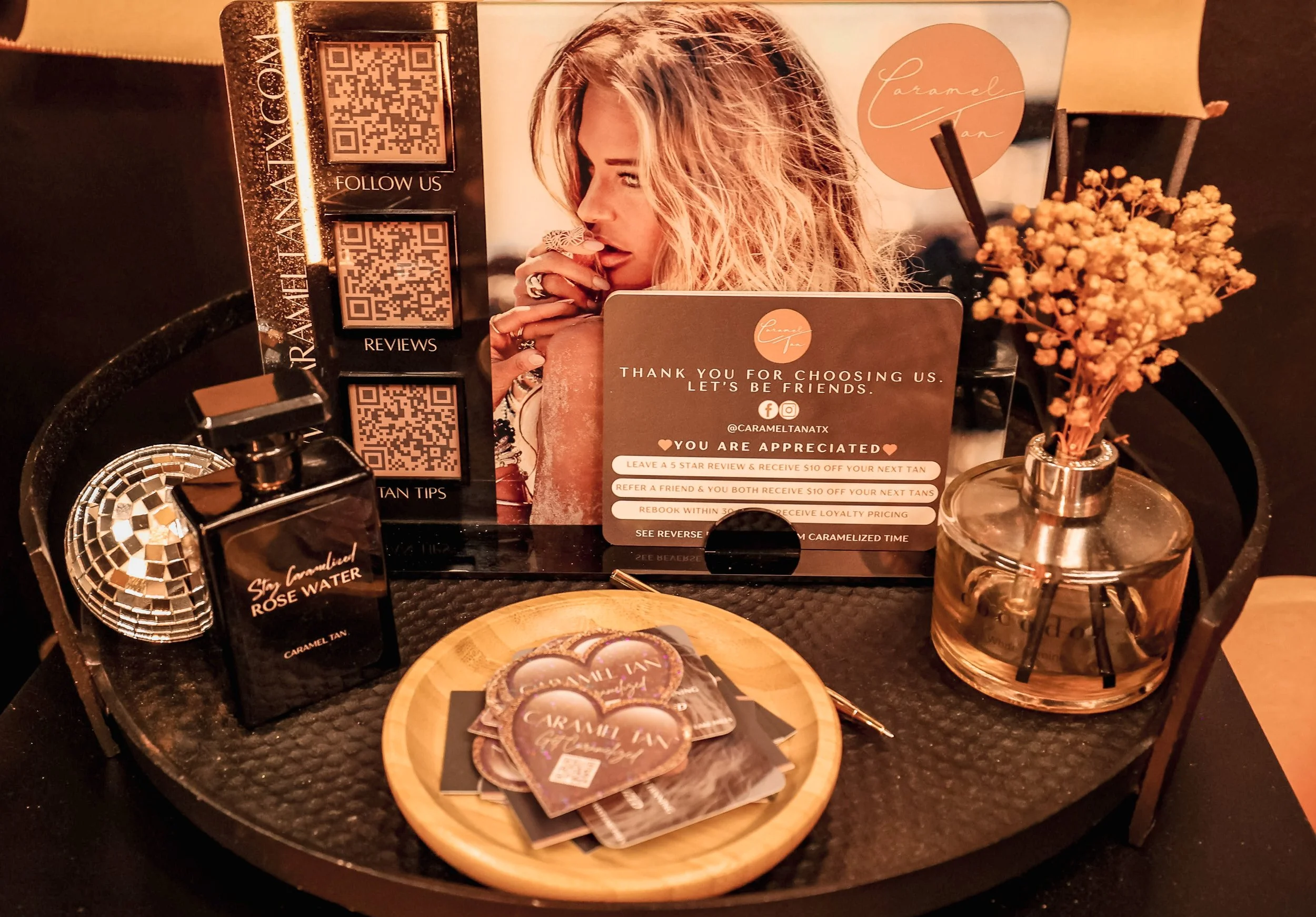 A promotional display on a round black tray featuring perfume bottles, business cards, a pen, a small bouquet of dried flowers in a glass bottle, and signs with QR codes and social media handles for a brand called Caramel Tan, specializing in caramelized tanning products.