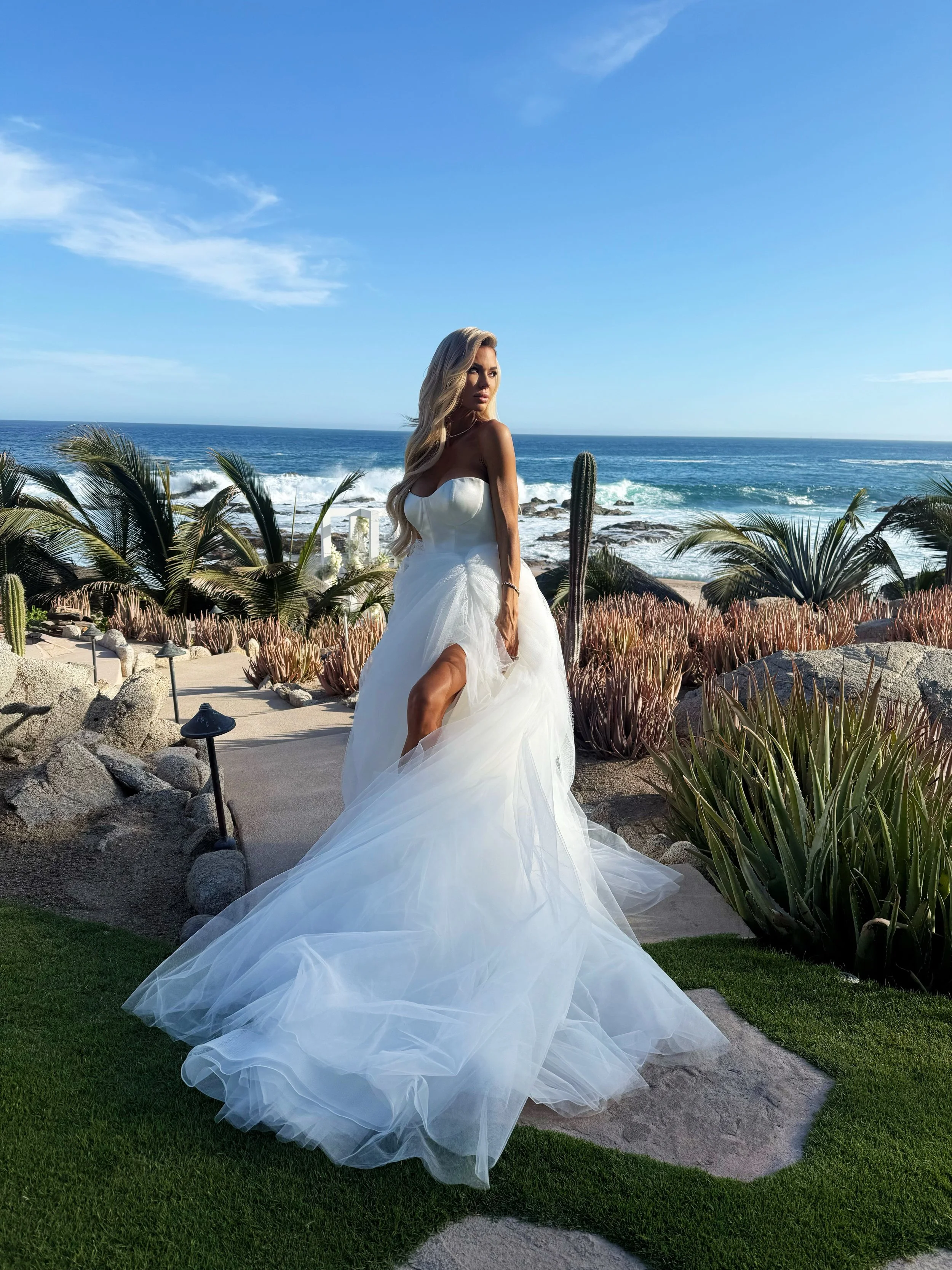 A bride with a gorgeous caramel tan in a white wedding dress standing on a pathway near the ocean with a clear blue sky, ocean waves, cacti, and various desert plants in the background.