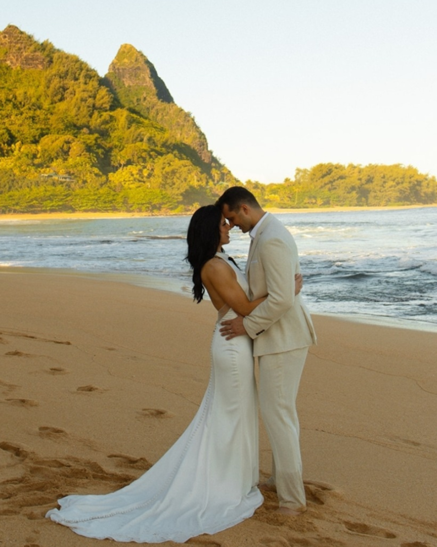 A couple dressed in wedding attire standing close together on a sandy beach with a mountain and ocean in the background, during sunset.