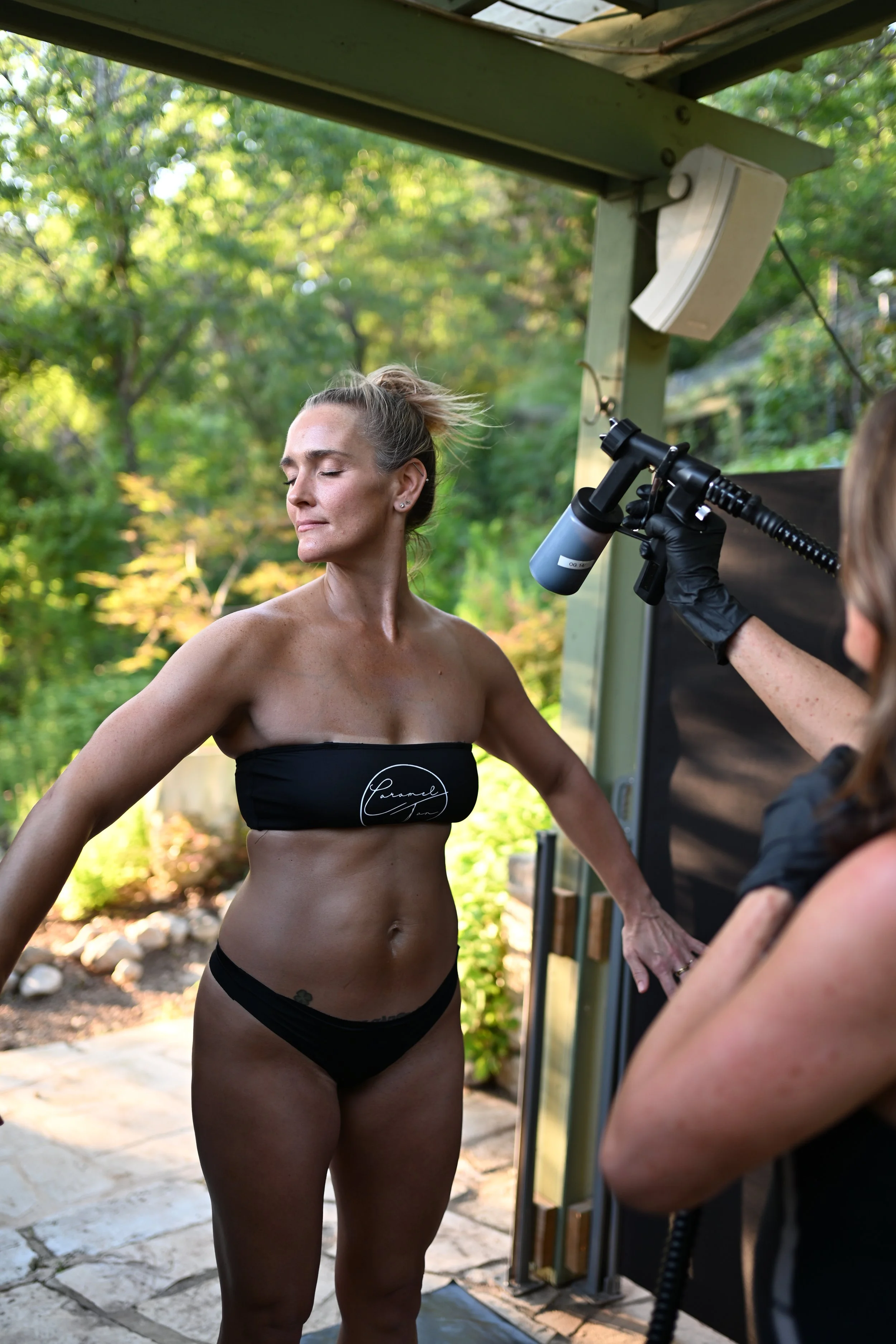 Spray tan artist applying a custom airbrush tan at the Caramel Tan Garden Studio, using clean, skin-loving formulas in a natural outdoor setting.