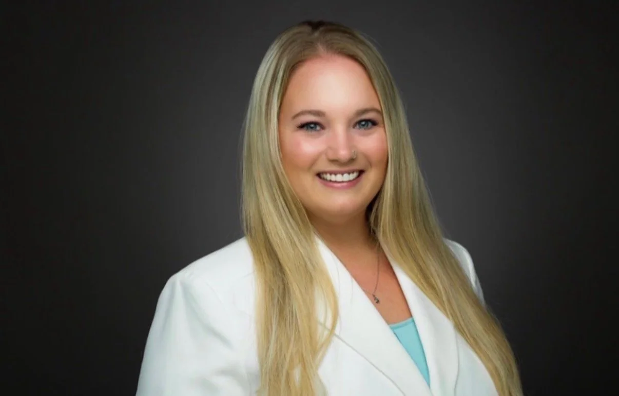 A young woman with long blonde hair wearing a white blazer and light blue top, smiling against a dark gray background.