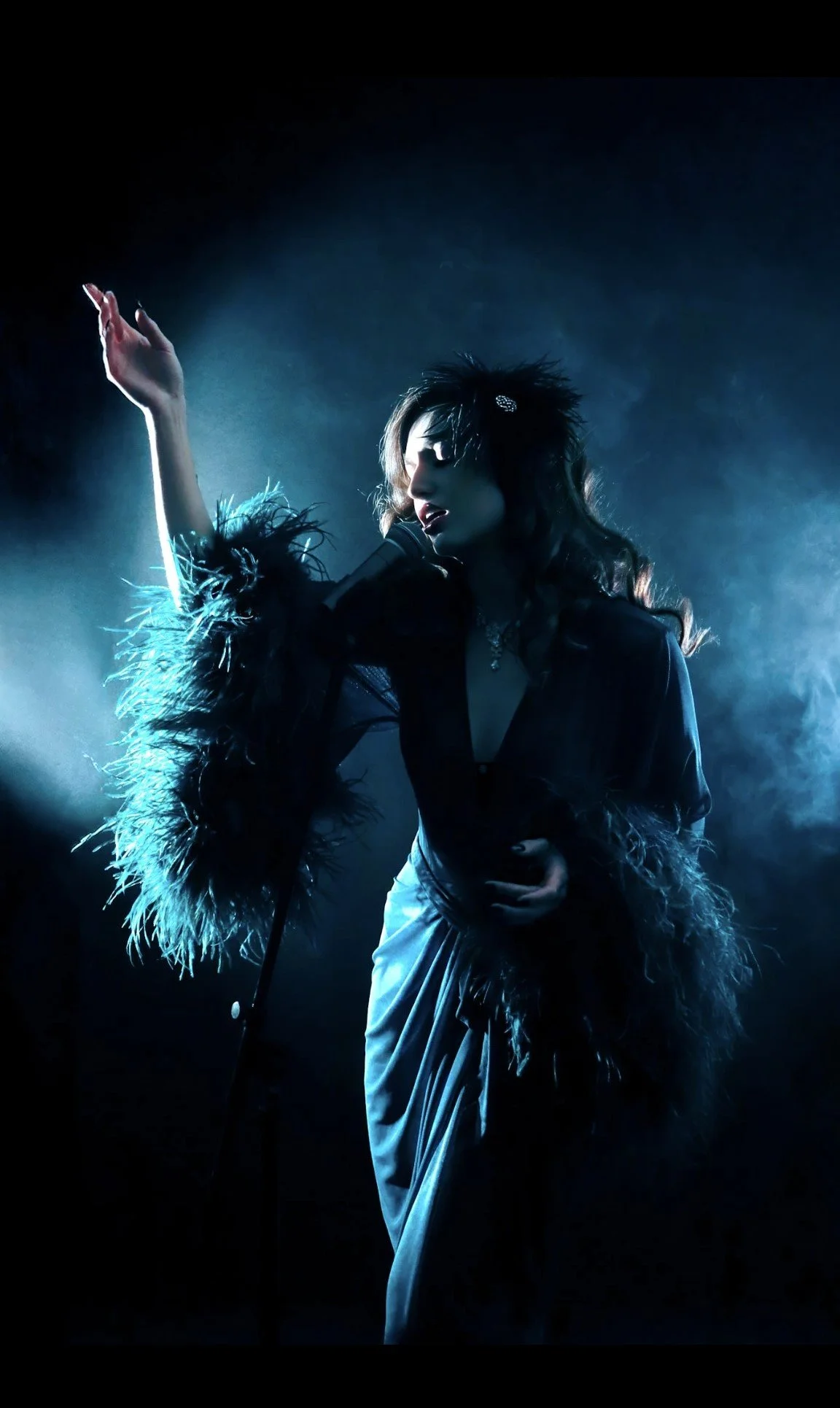 A female singer performing on stage with dramatic lighting and smoky background, wearing a dark outfit with feathered details.