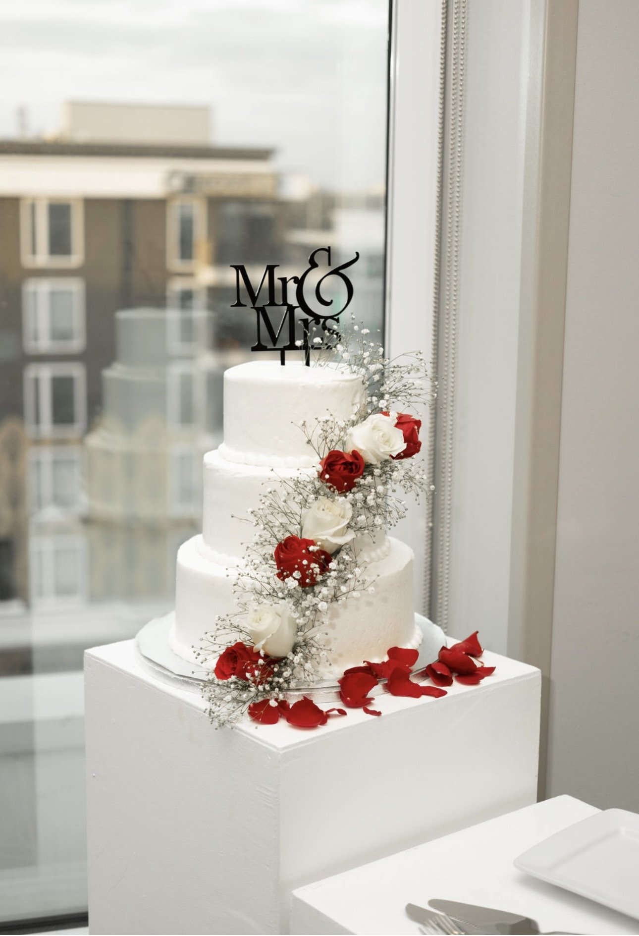 A three-tier white wedding cake decorated with red and white roses and baby's breath, topped with a 'Mr & Mrs' cake topper, placed on a white pedestal near a window.