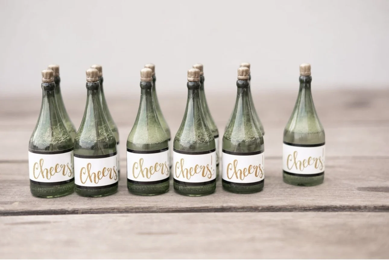 Six miniature glass bottles with cork stoppers, each labeled 'Cheers' in gold lettering, arranged in two rows on a wooden surface.
