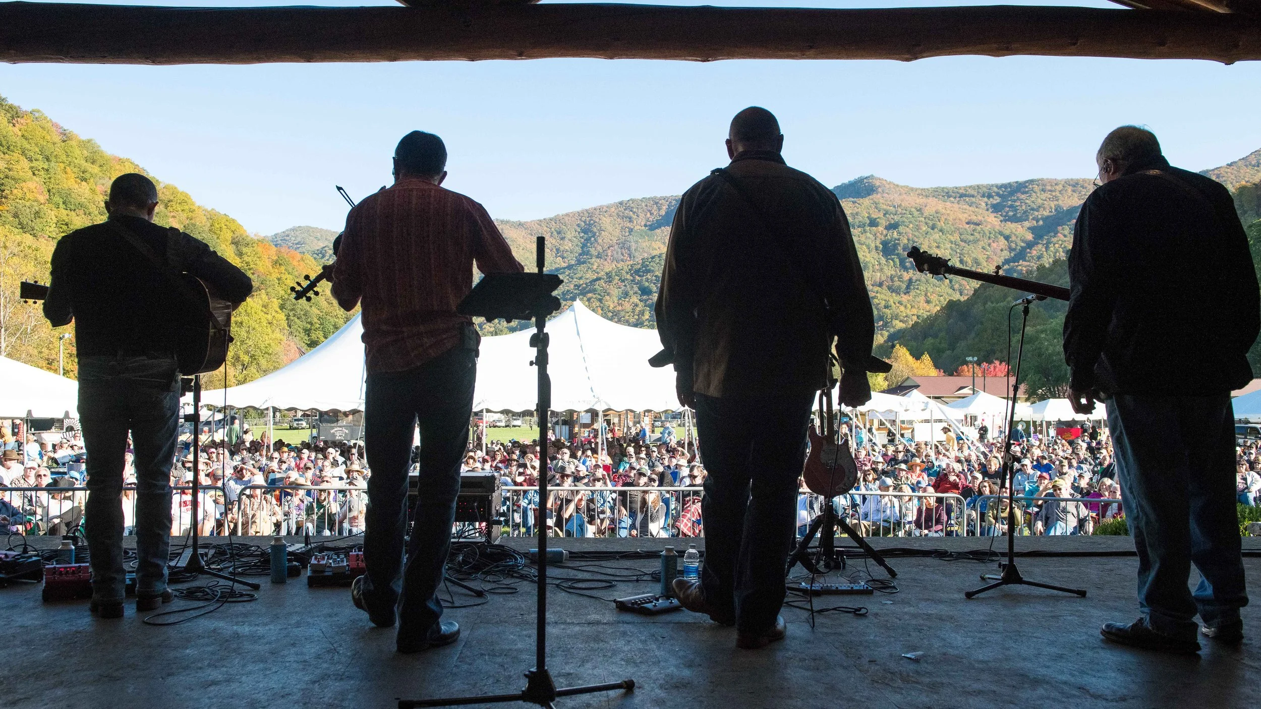 View of a live outdoor musical performance with four musicians on stage, overlooking a crowd at a festival in a mountainous area during daytime.