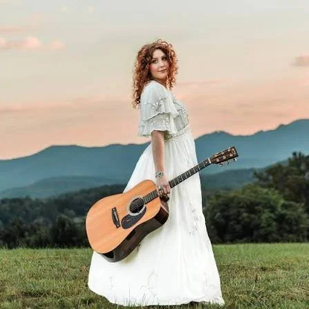 A woman in a white dress holding an acoustic guitar outdoors with mountains and a colorful sky in the background.