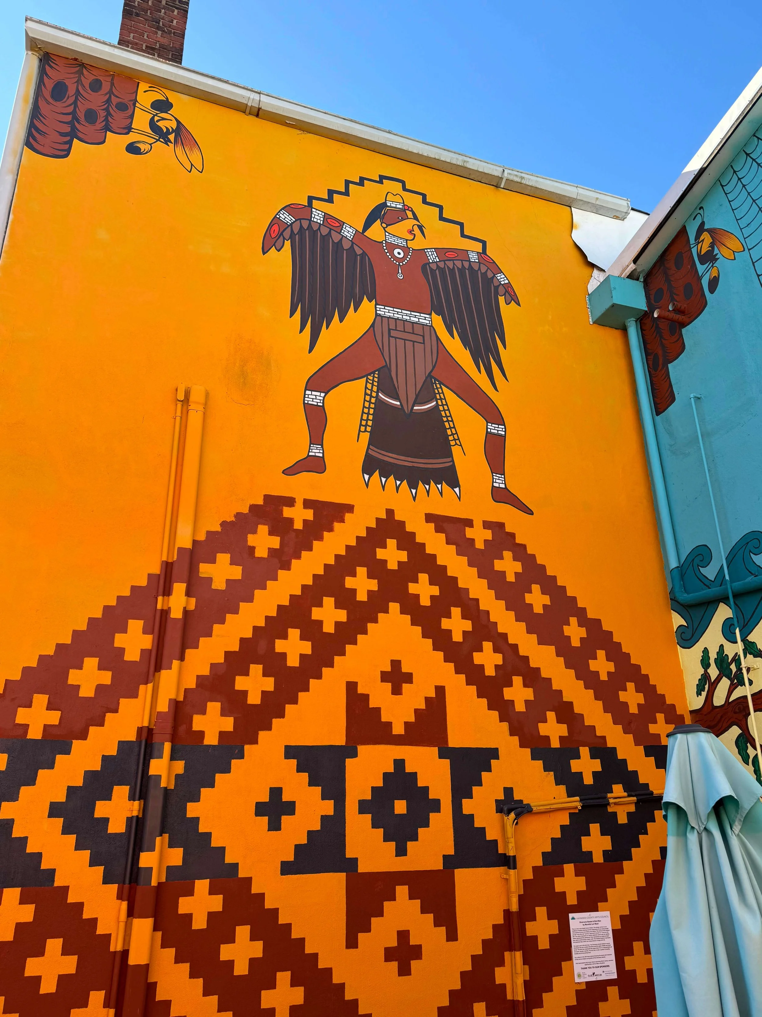 Colorful mural of a stylized bird with outstretched wings, wearing jewelry and a headdress, against a bright yellow background, with a geometric patterned skirt below.