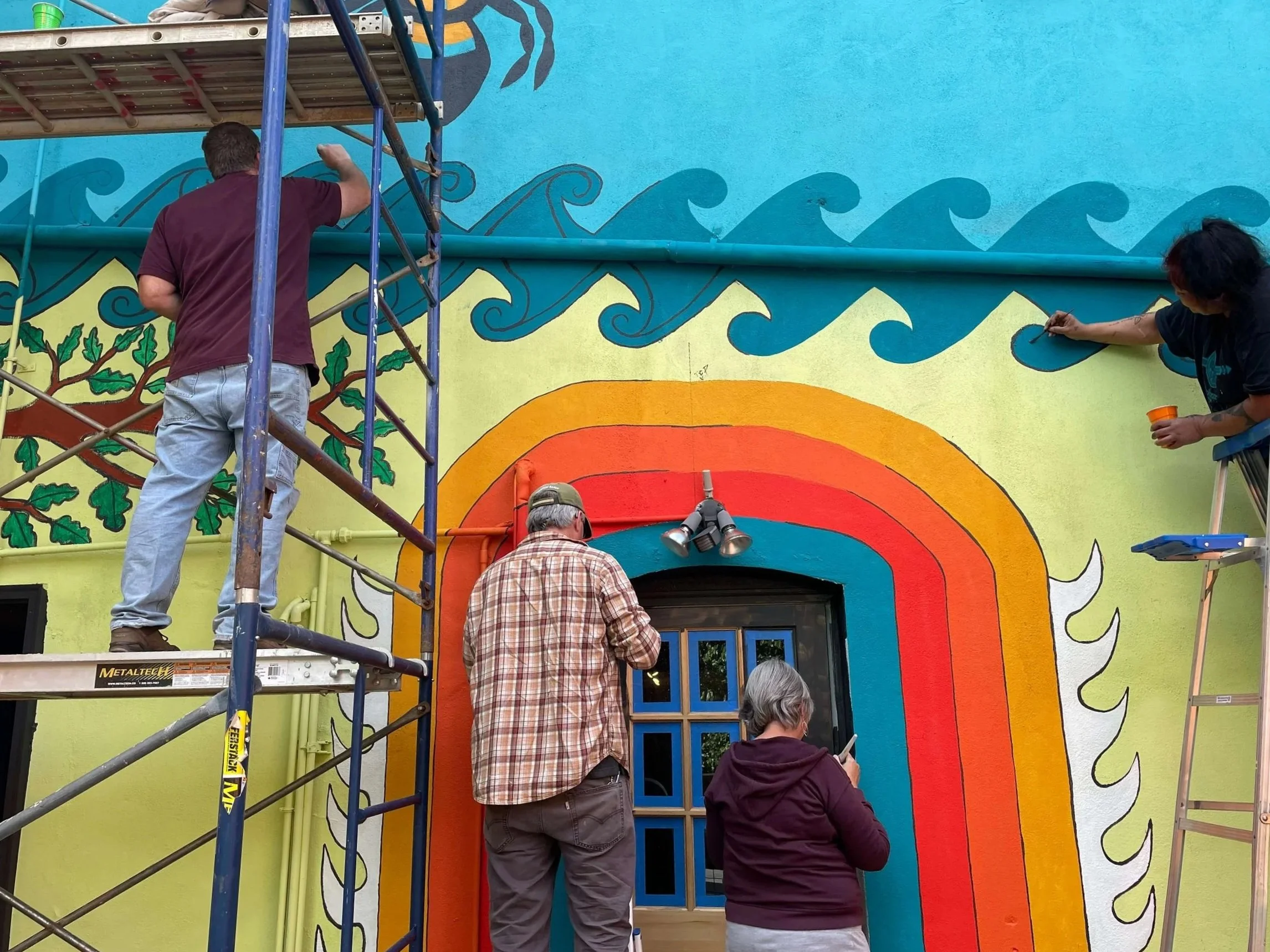 People painting a colorful mural on the exterior wall of a building. The mural features waves, a rainbow, a tree, and other vibrant elements. Scaffolding is set up to assist the artists.