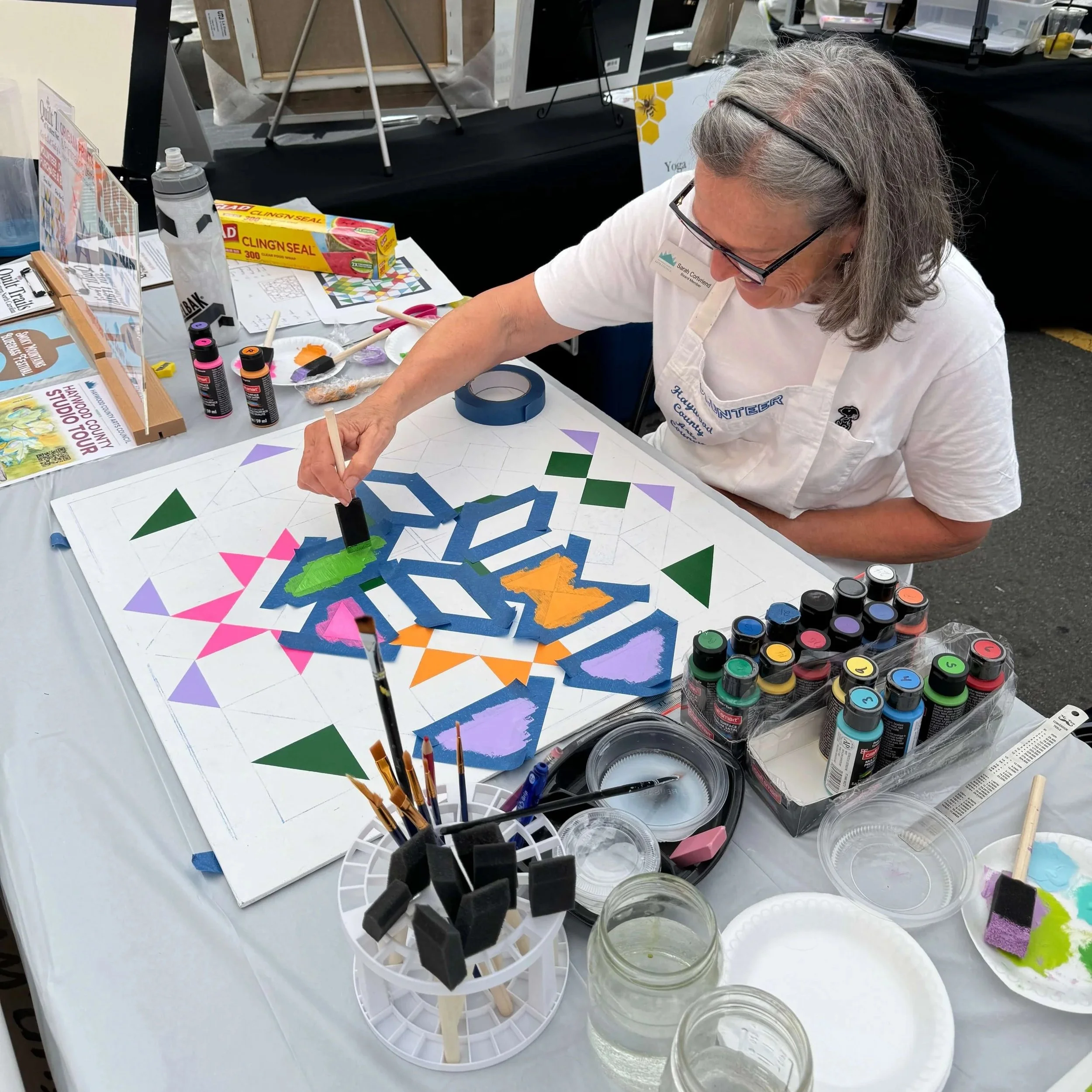 A woman working on a geometric art project at a craft booth, with various paints, brushes, and craft supplies on the table.