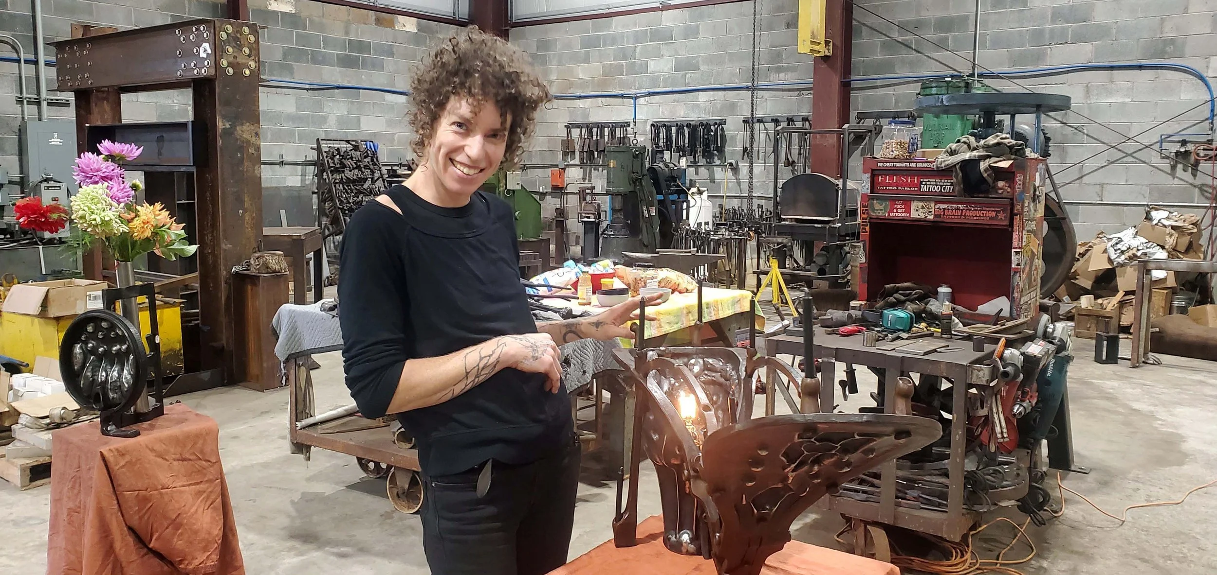 A woman with curly hair and tattoos smiling in a woodworking or metalworking workshop, standing next to a table with a decorative metal object and a flower arrangement in the background.