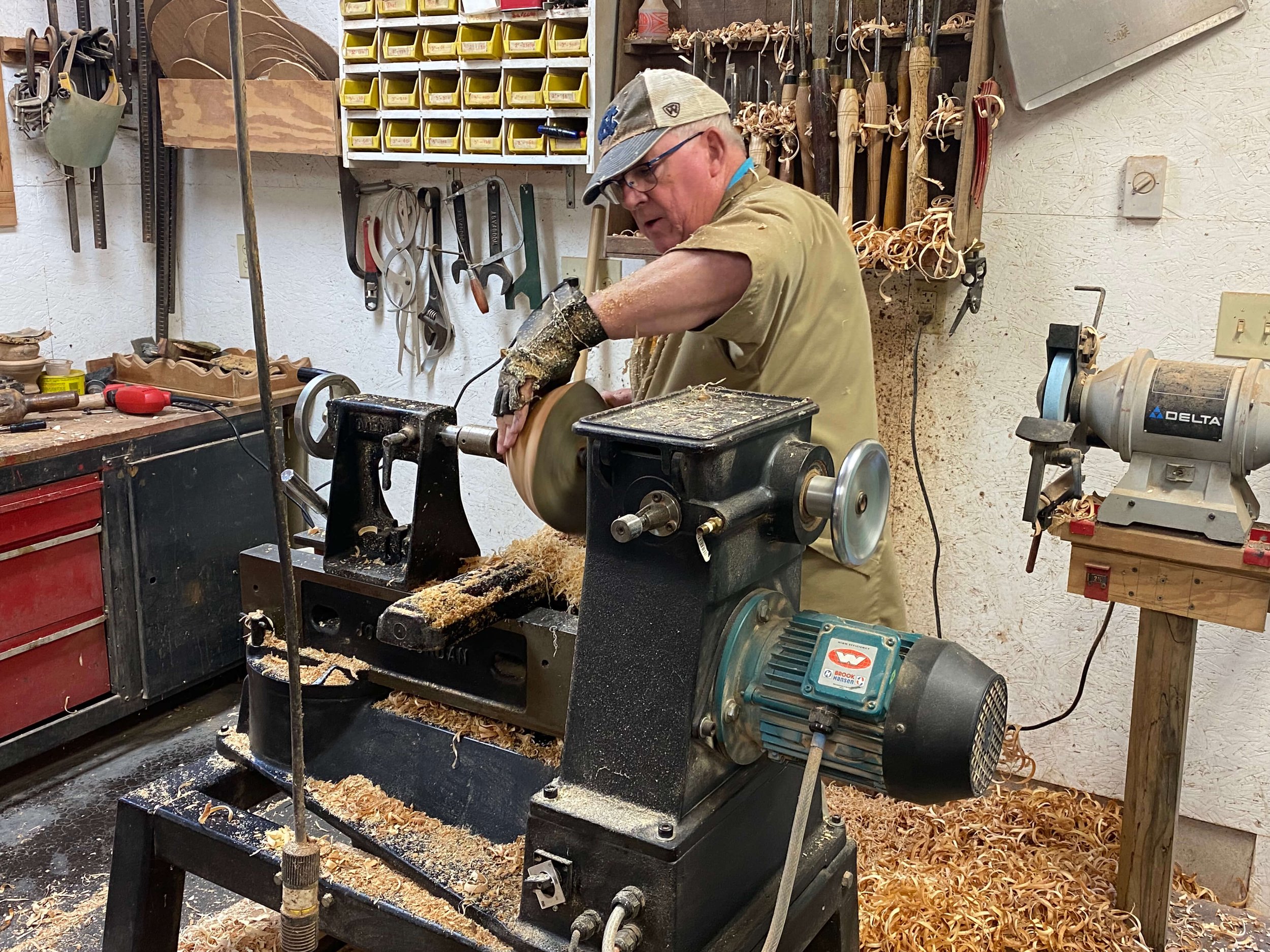 A man wearing glasses, a cap, and a beige shirt operating a wood lathe in a woodworking shop. The shop has various woodworking tools hanging on the wall and shelves with small storage bins.