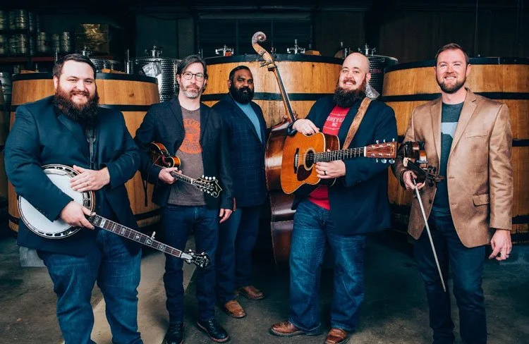 Five men holding musical instruments, standing in front of a bar with wooden tanks, smiling at the camera.