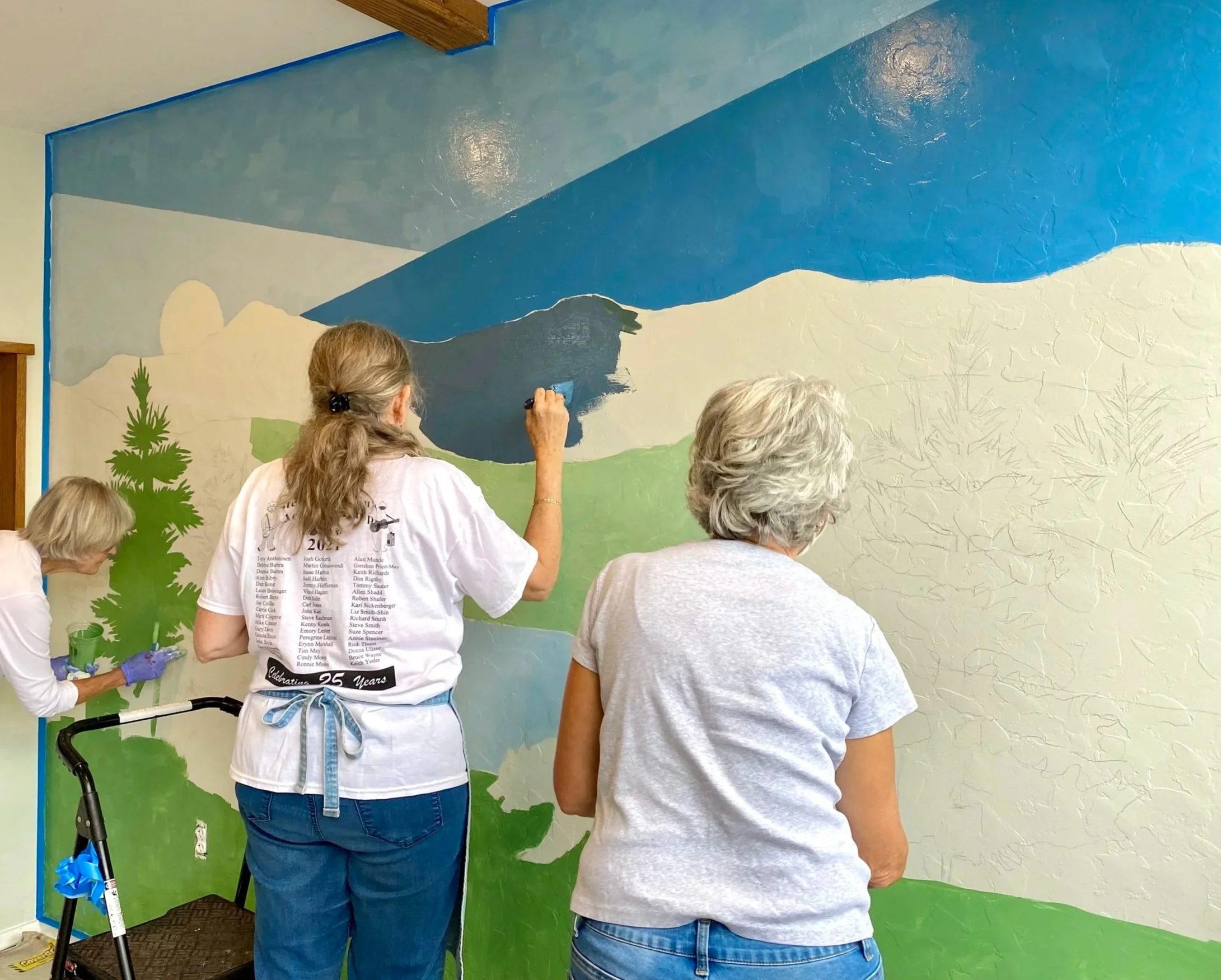 People painting a mural on a wall with a mountain scene, some individuals are seen from the back.