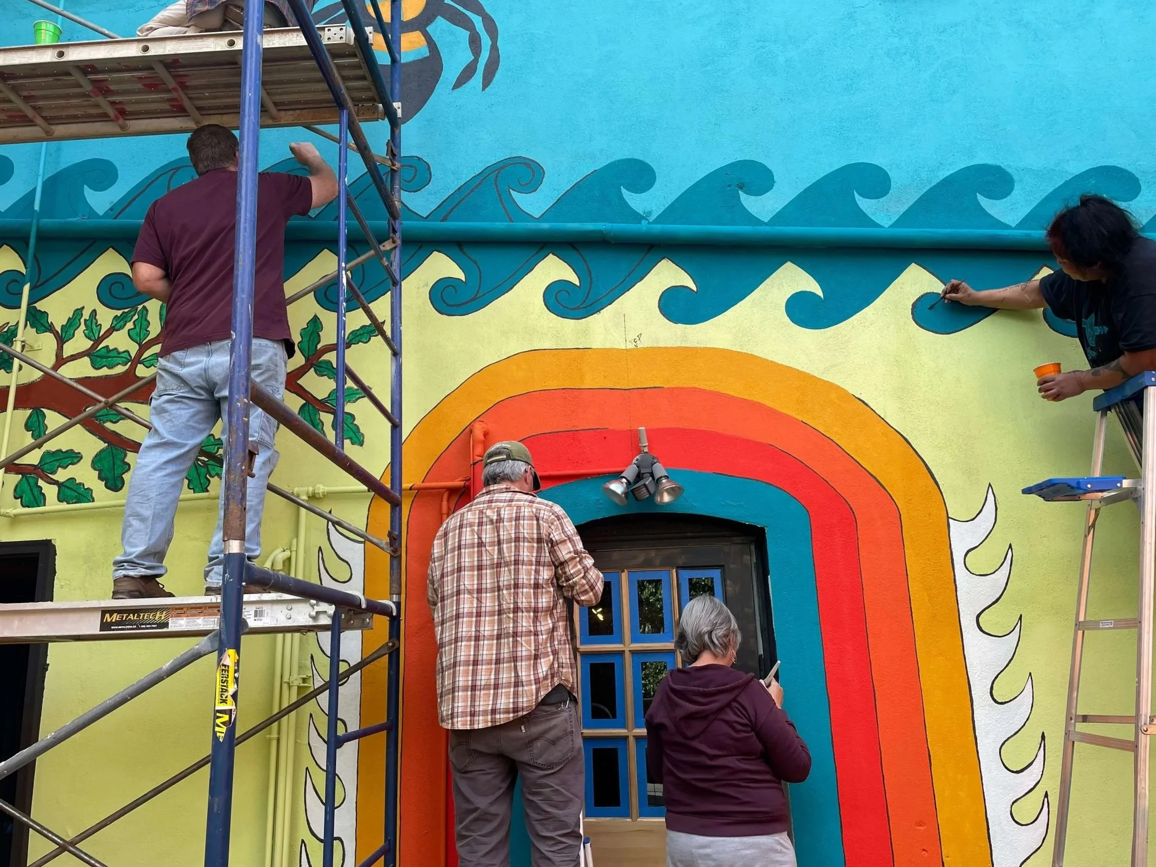 People painting a colorful mural on a building wall, featuring a rainbow, waves, and a tree with leaves, with scaffolding and tools around them.