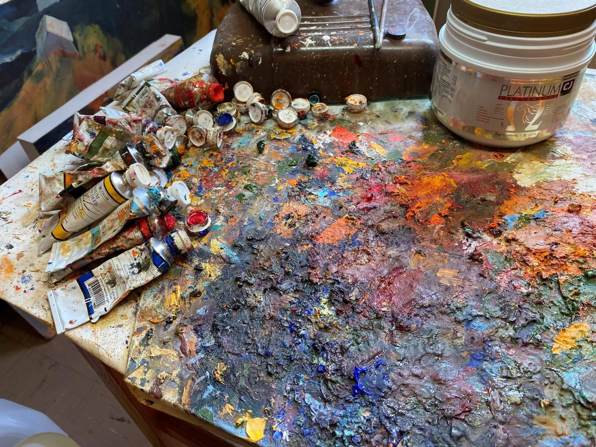 A cluttered artist's workspace with a table covered in colorful paints, paint tubes, and paint caps. There is a wooden or metal container on the table, and containers of white paint or modeling paste. The workspace is messy, with dried and spilled paint across the surface.