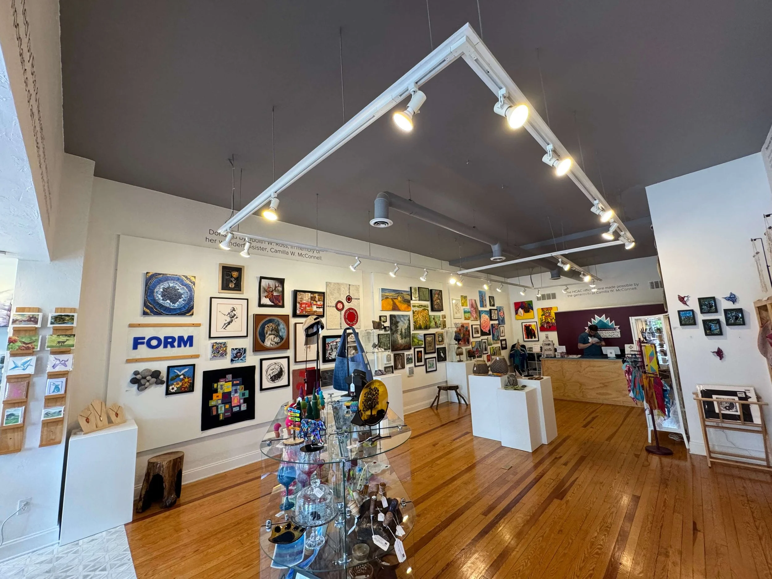 An art gallery showcasing various paintings, sculptures, and decorative items on display walls and tables, with gallery lighting illuminating the artwork.
