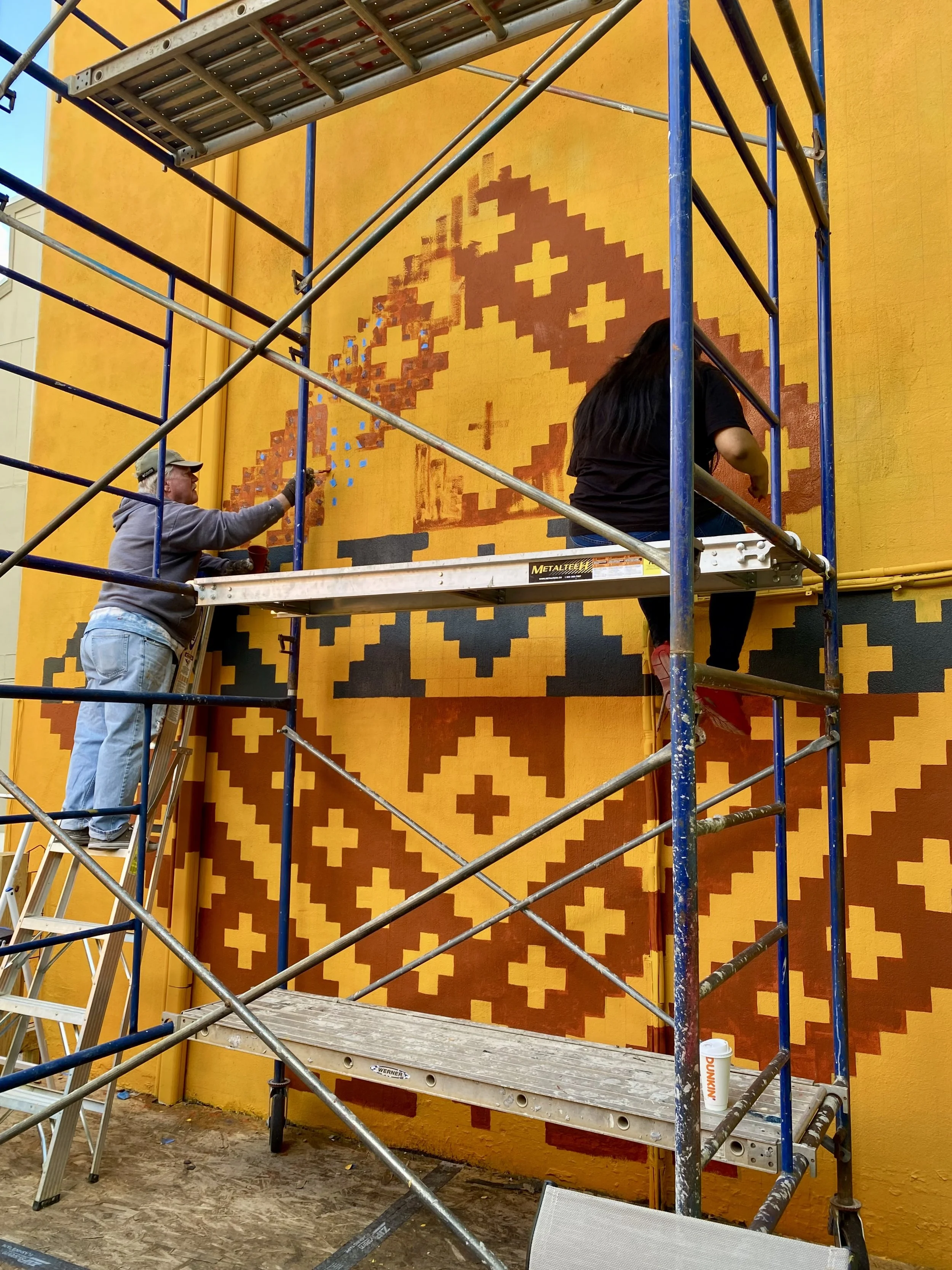 Two artists working on a pixelated mural of a character with a yellow face and darker features on a yellow wall, surrounded by scaffolding.