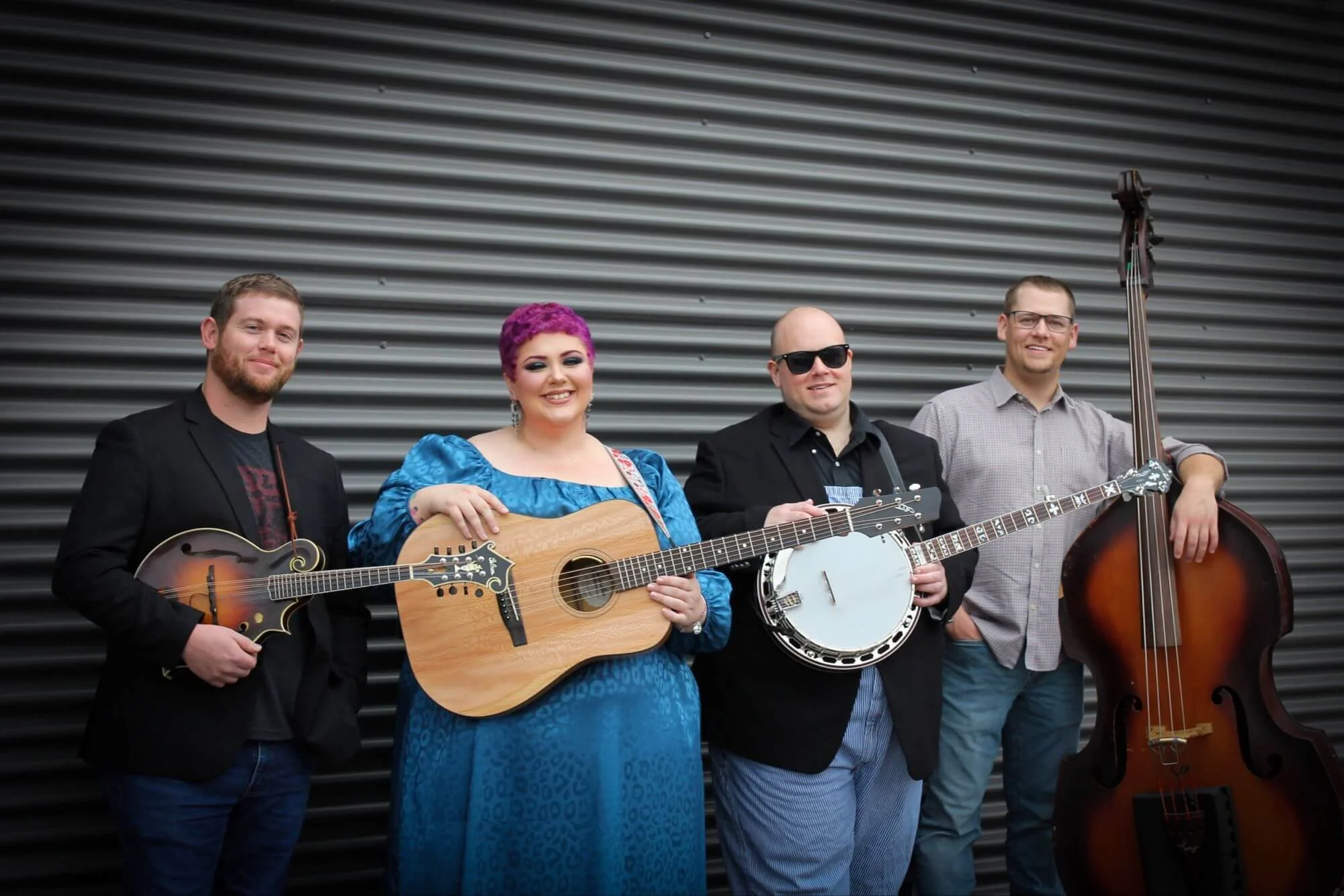 A group of four musicians standing against a corrugated metal wall, holding acoustic and electric string instruments, posing for a photo and smiling.