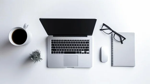 Top view of a workspace with a coffee cup, a laptop, eyeglasses, a computer mouse, a notepad, and a small plant on a white surface.