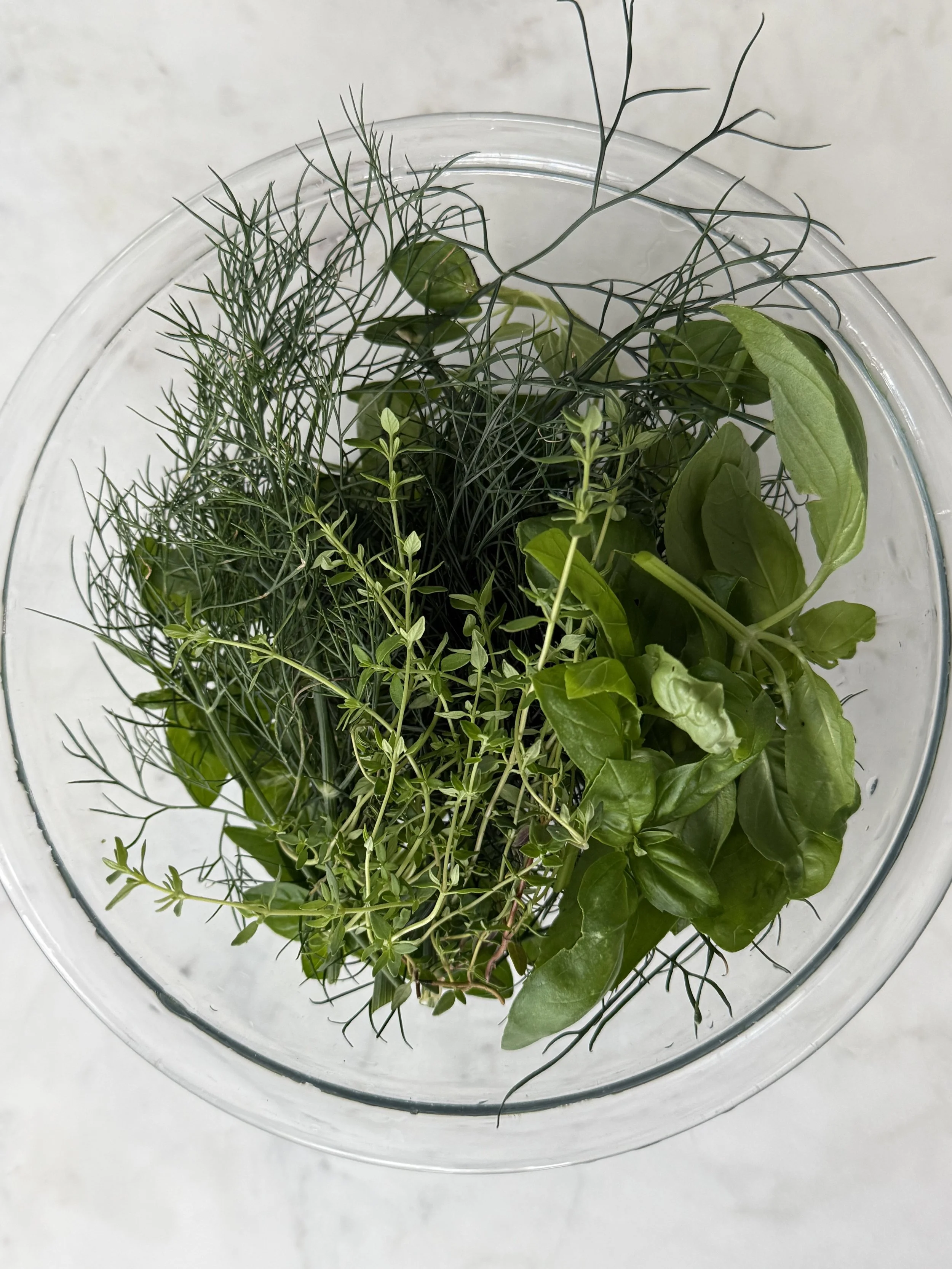 Top view of a clear bowl filled with a variety of fresh green herbs including basil, thyme, and dill, on a white surface.
