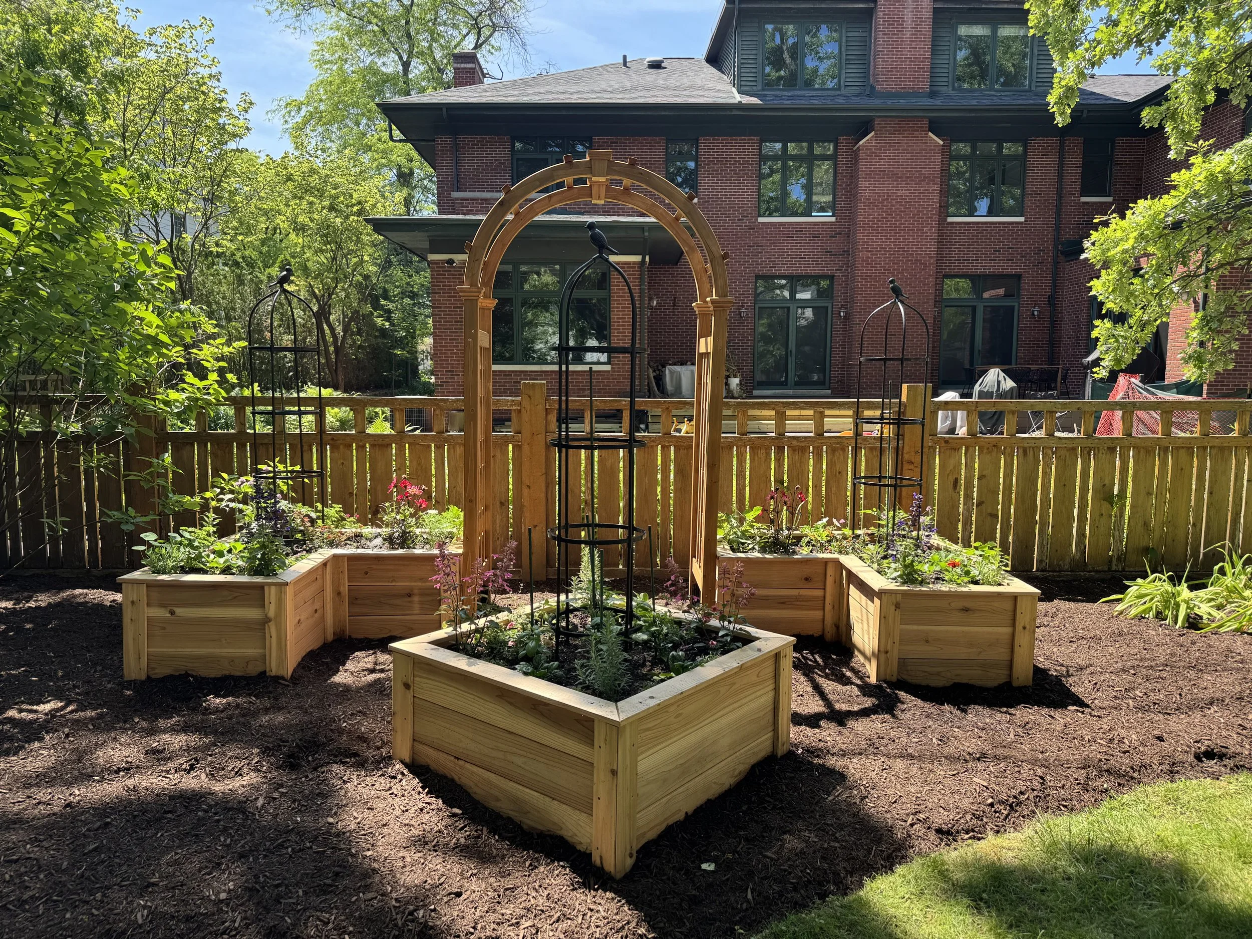A backyard garden with raised wooden beds planting various flowers and vegetables, surrounded by a wooden fence, with a brick house in the background and trees providing shade.